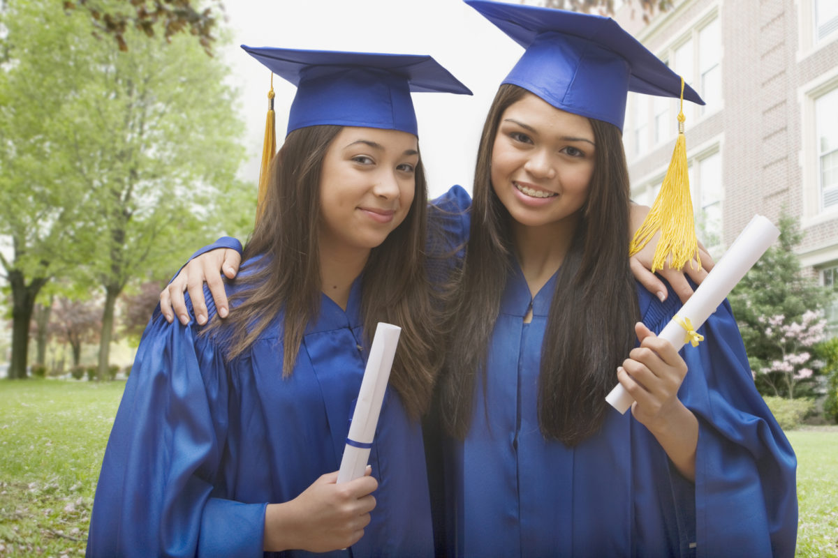 Multi-ethnic girls in caps and gowns holding diplomas and hugging coaching